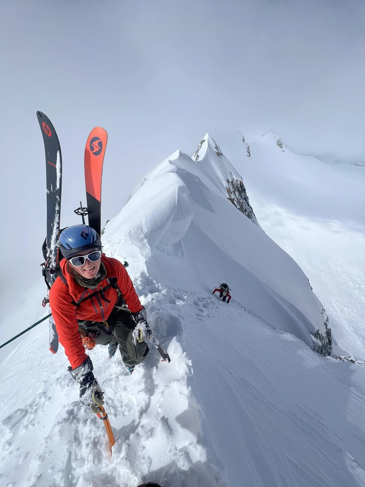 A smiling climber carrying skis along a snowy ridge