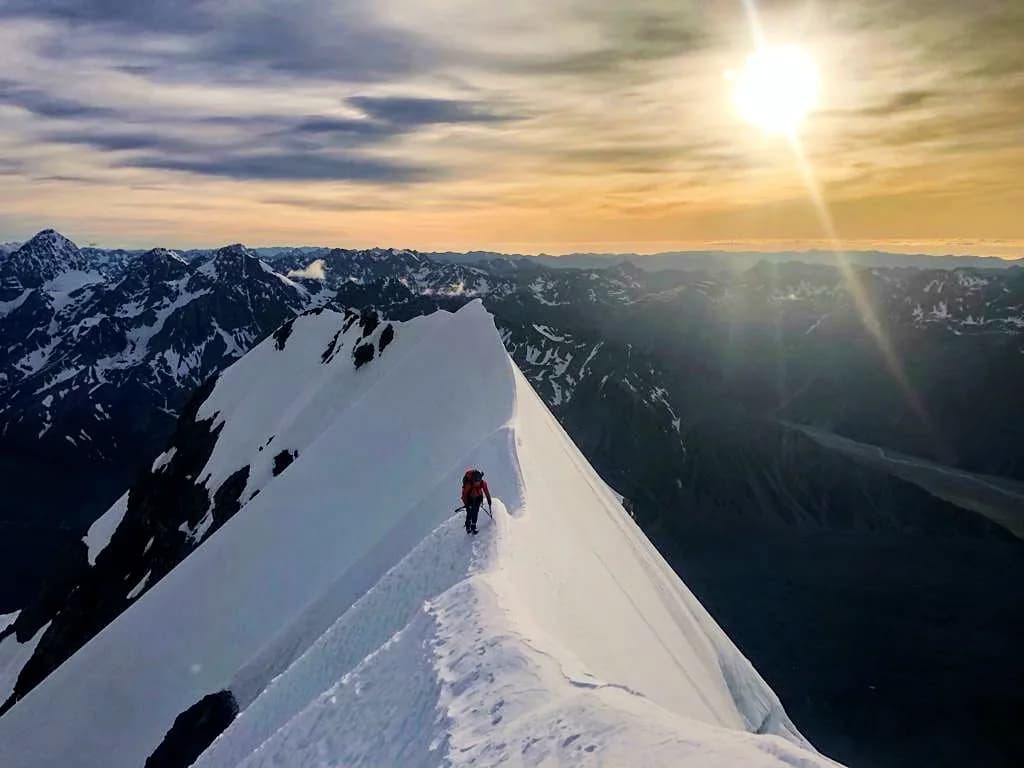 A mountaineer on a sunlit snow ridge at first light, with alpine peaks beyond