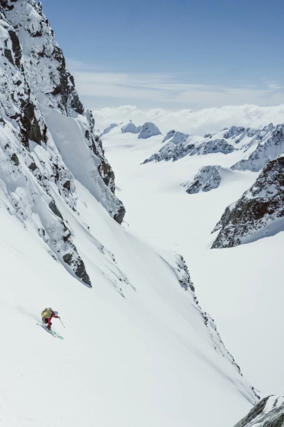 A skier in a red jacket descending a steep snow slope above a wide alpine basin